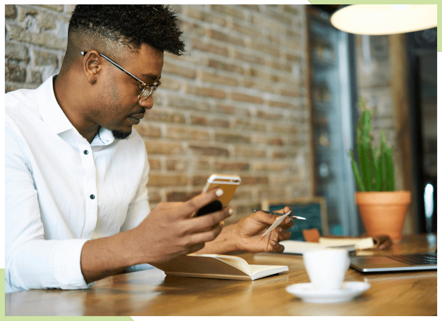 Man sitting down at a table with coffee holding his phone with a book open next to his laptop