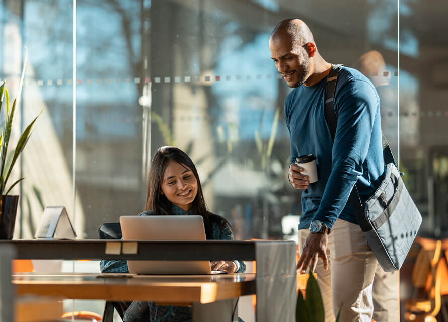 Female sitting at desk with laptop while man stands and chats besides her with a coffee