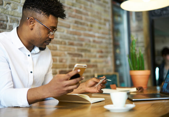 Man sitting down at a table with coffee holding his phone with a book open next to his laptop