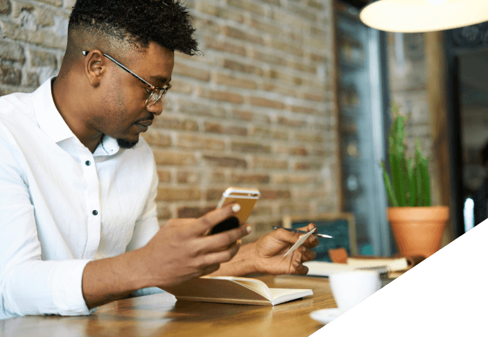 Man sitting down at a table with coffee holding his phone with a book open next to his laptop