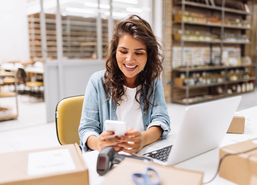 Female small business owner sitting at desk holding her phone next to a laptop