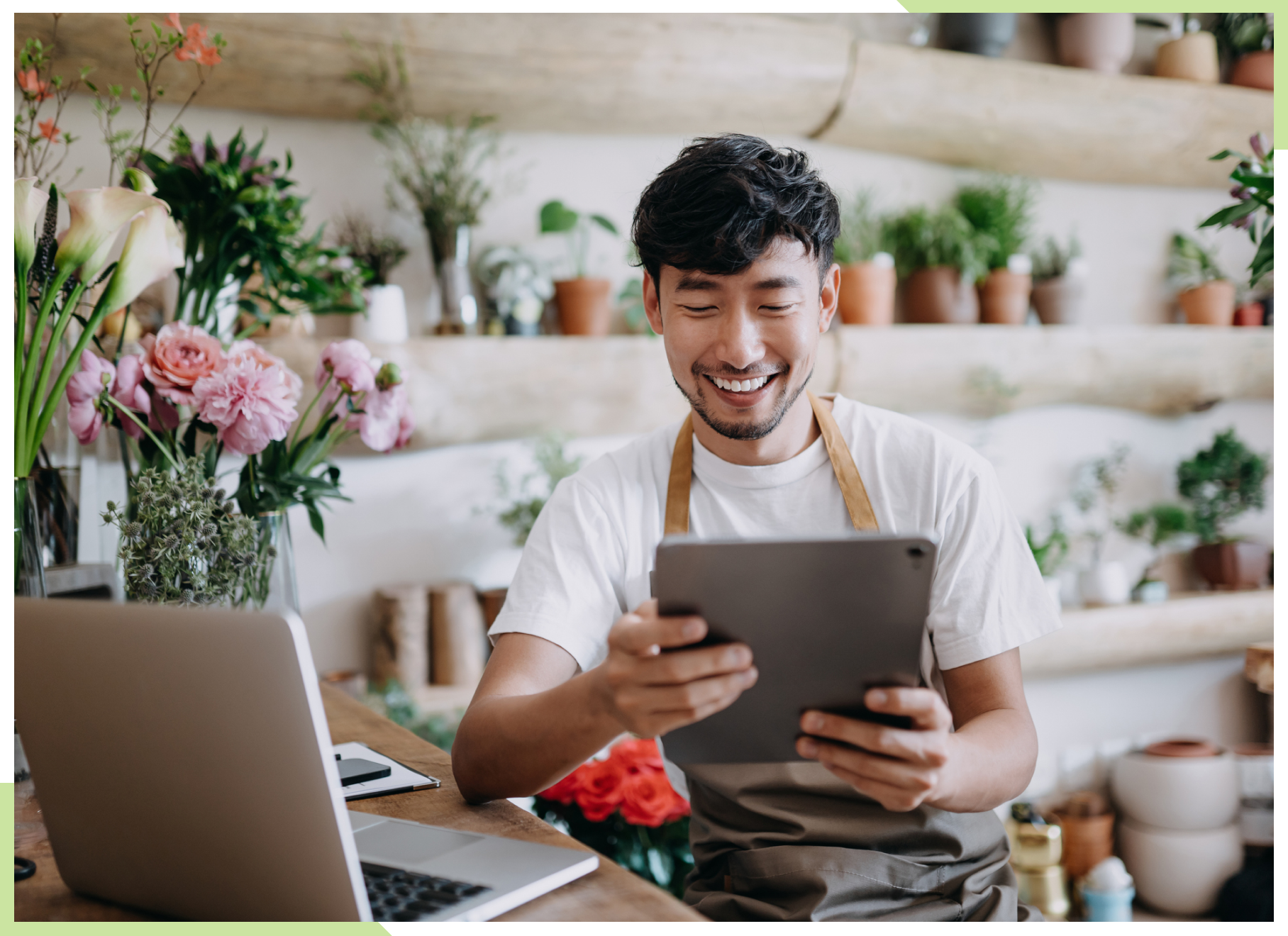 A man working in a flower shop performs financial services for his business, including domestic wires, on his laptop while holding a tablet.