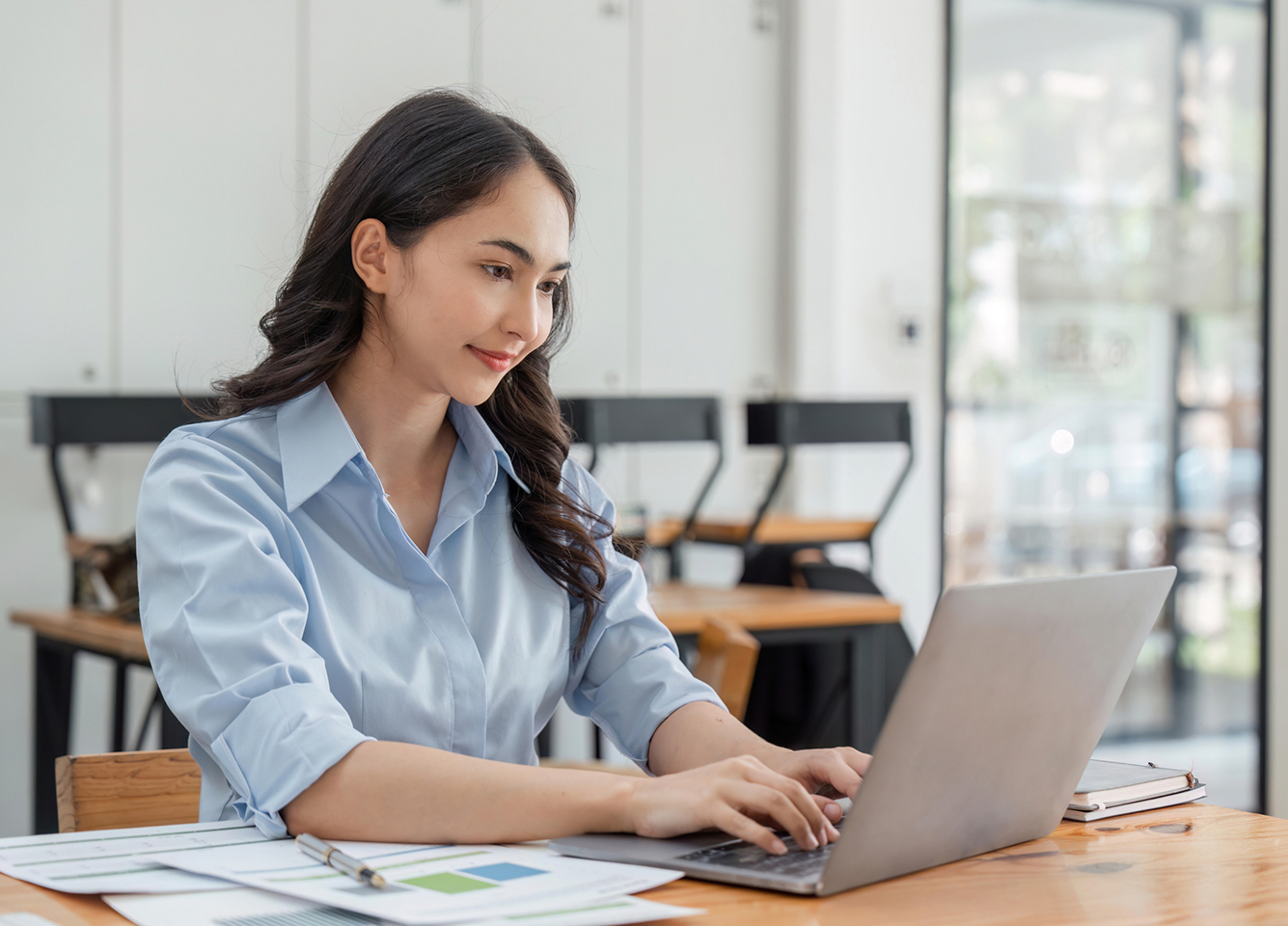 A woman in a small office performs financial transactions on a laptop with reports on the desk beside her.